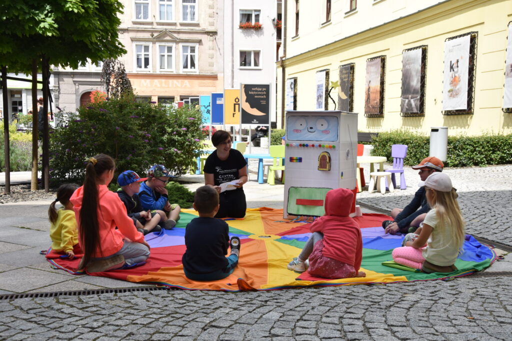 Góra bajek - spotkanie z Bajkomatem, dzieci siedzące na kolorowej chuście słuchające pani bibliotekarki