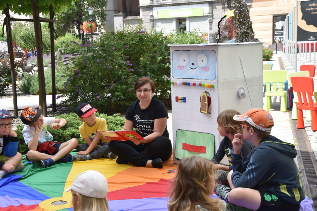Góra bajek - spotkanie z Bajkomatem, dzieci siedzące na kolorowej chuście słuchające pani bibliotekarki