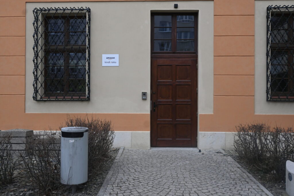 Boczne wejście do budynku z drewnianymi drzwiami, oknami zabezpieczonymi kratami oraz przy kamiennej ścieżce.