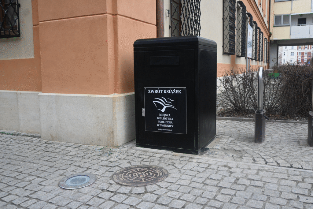 Czarna skrzynia z napisem zwrot książek, logiem i nazwą Miejska Biblioteka Publiczna w Świdnicy. Jest przy chodniku obok budynku.