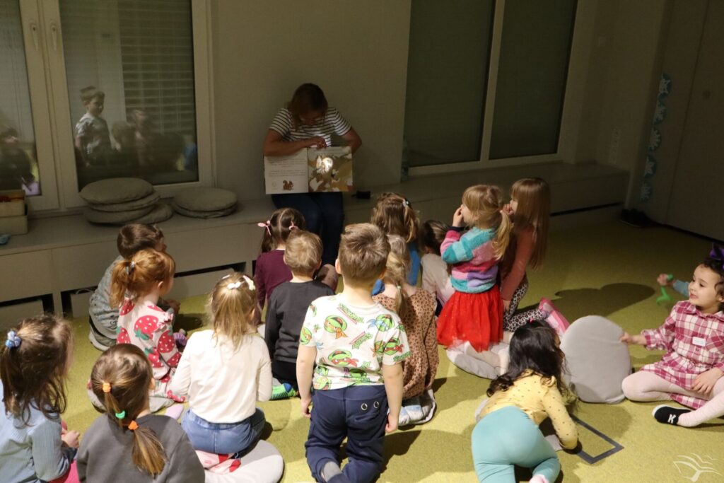 Prowadząca świeci latarką na książkę. Dzieci zwrócone do niej obserwują książkę.