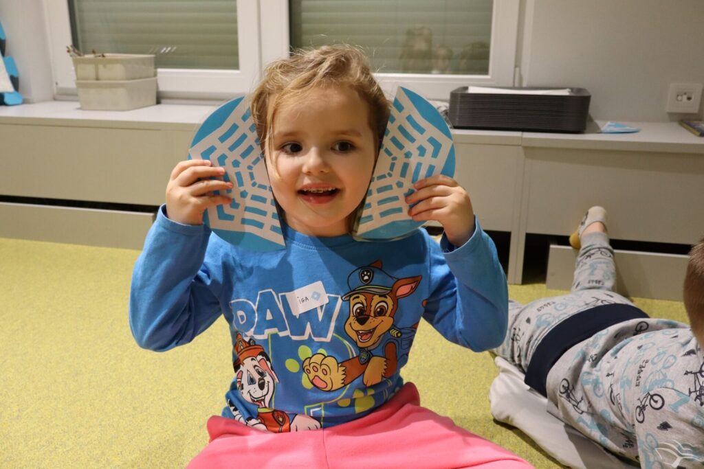 Dziewczynka siedzi i trzyma obok głowy dwie połowy śnieżynki zrobionej z papieru.