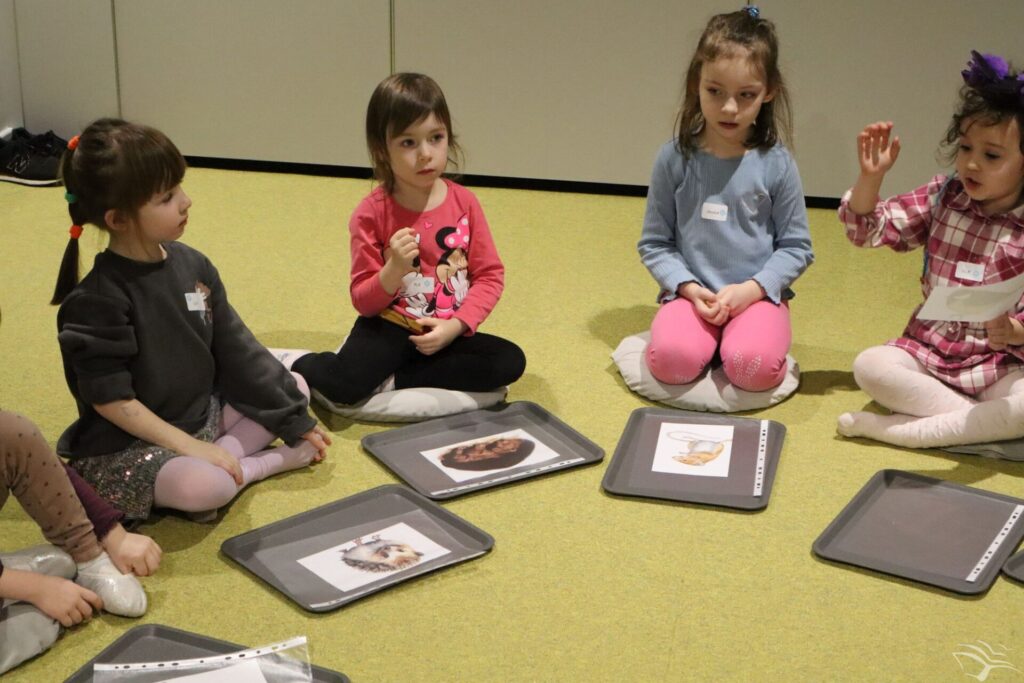 Dzieci siedzą dywanie. Przed nimi znajdują się rysunki.
