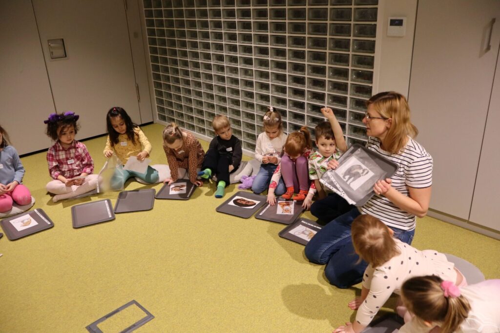 Dzieci i bibliotekarka siedzą na dywanie i oglądają rysunki.